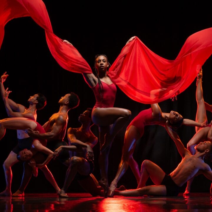 A group of dancers pose dramatically on stage, with one dancer in the center holding flowing red fabric above her head.