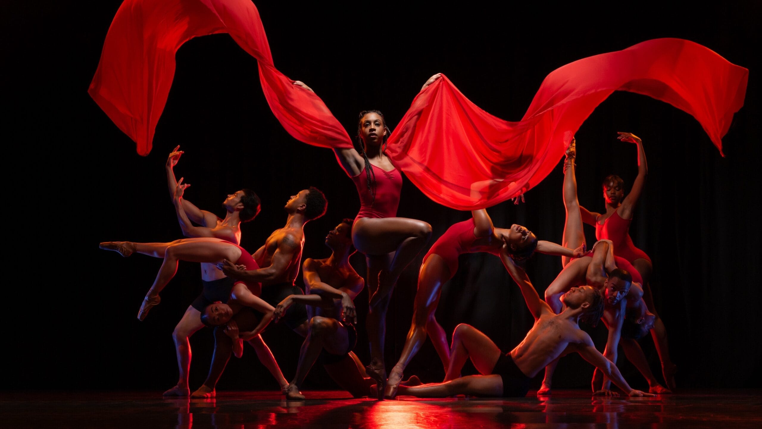 A group of dancers pose dramatically on stage, with one dancer in the center holding flowing red fabric above her head.