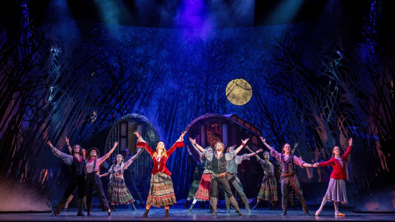 A group of actors in colorful period costumes perform a synchronized dance on stage with a forest backdrop and a full moon.