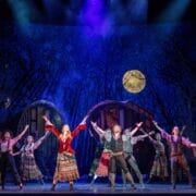A group of actors in colorful period costumes perform a synchronized dance on stage with a forest backdrop and a full moon.