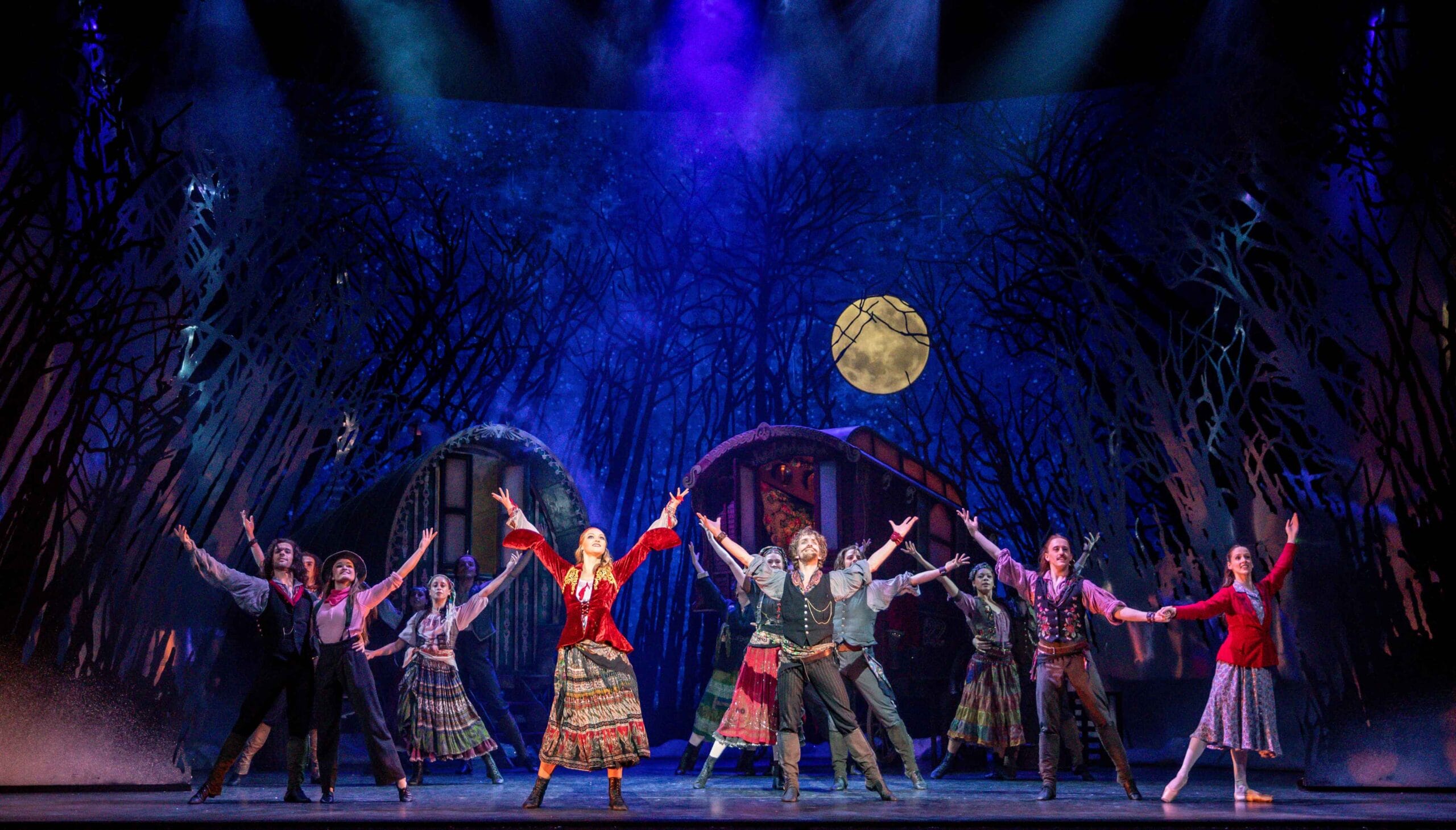A group of actors in colorful period costumes perform a synchronized dance on stage with a forest backdrop and a full moon.