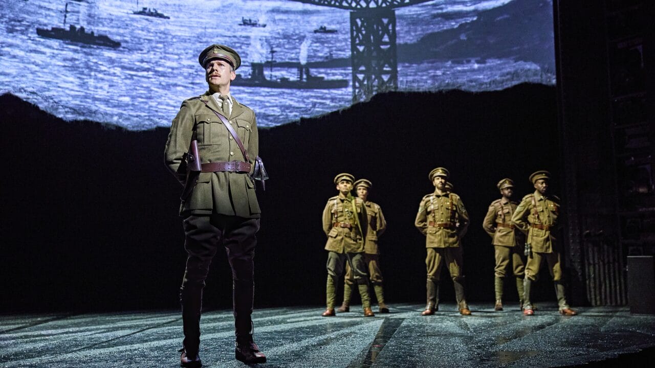Six actors in military uniforms stand on stage with a backdrop depicting ships and a crane, suggesting a wartime setting.