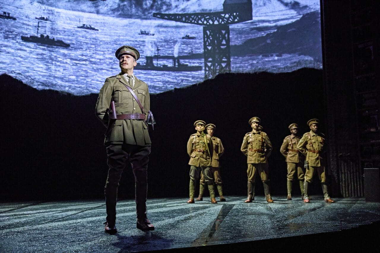 Six actors in military uniforms stand on stage with a backdrop depicting ships and a crane, suggesting a wartime setting.