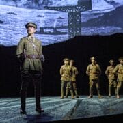 Six actors in military uniforms stand on stage with a backdrop depicting ships and a crane, suggesting a wartime setting.