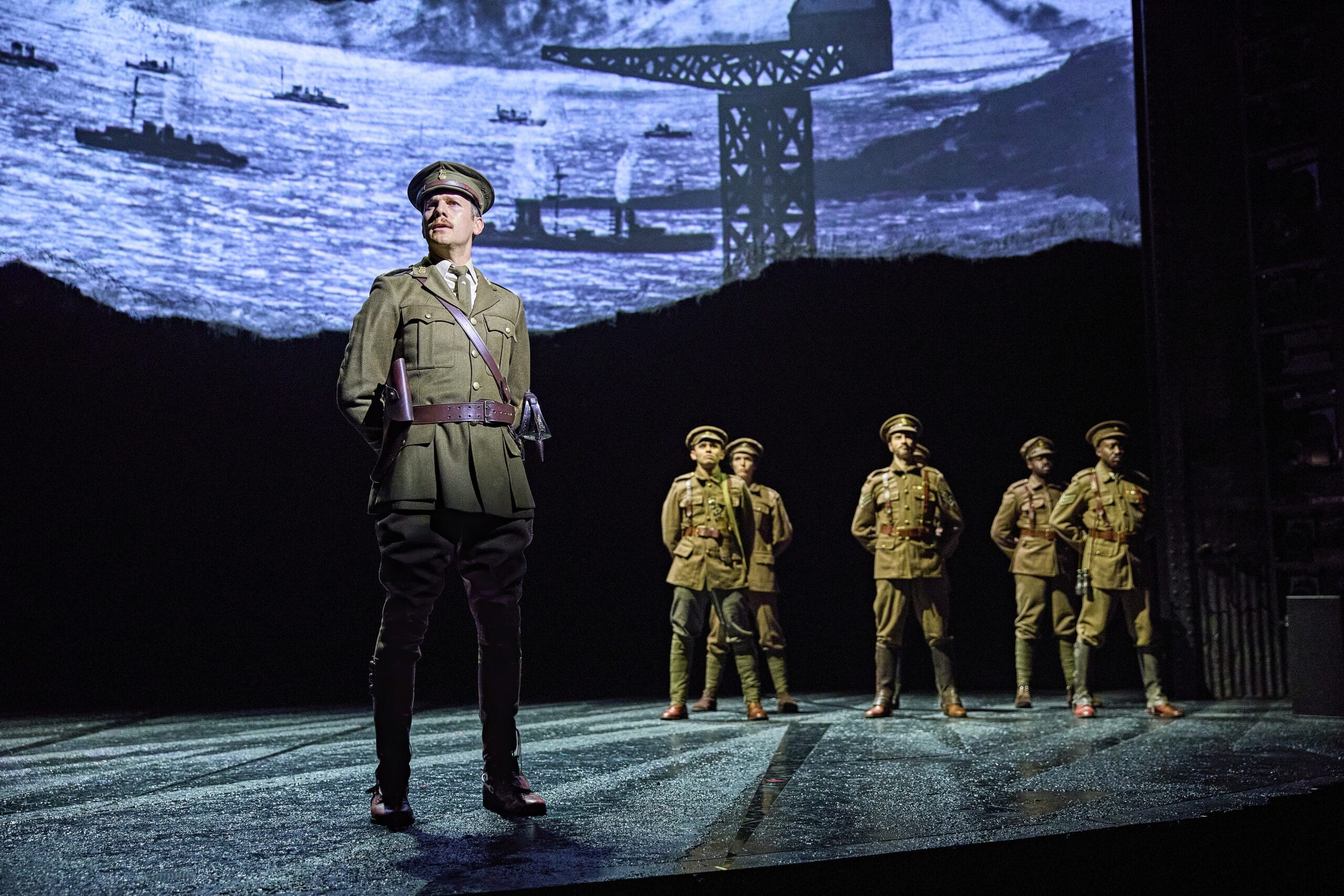 Six actors in military uniforms stand on stage with a backdrop depicting ships and a crane, suggesting a wartime setting.
