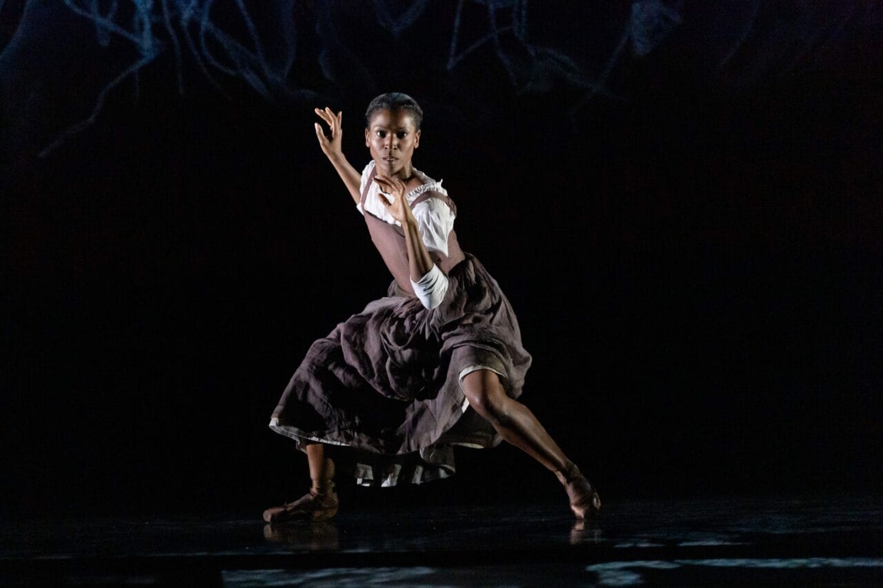 A dancer performs on stage, wearing a white top and a brown skirt, striking a dynamic pose with one arm raised and one leg extended against a dark background.