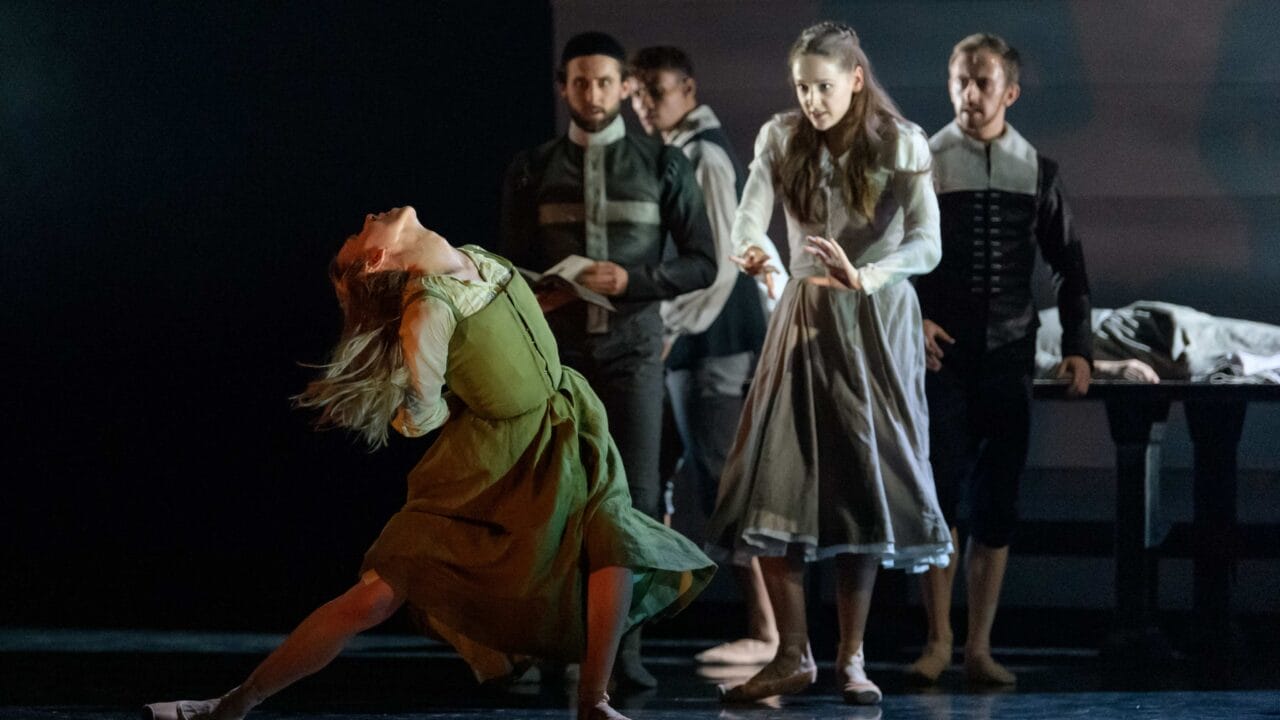 A group of dancers perform on stage; a woman in a green dress dramatically leans back while others in period costumes watch or gesture.