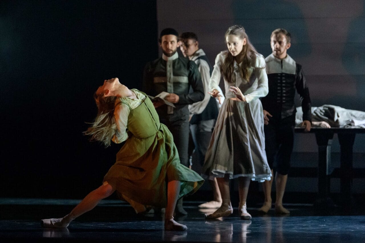 A group of dancers perform on stage; a woman in a green dress dramatically leans back while others in period costumes watch or gesture.