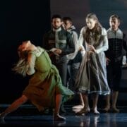 A group of dancers perform on stage; a woman in a green dress dramatically leans back while others in period costumes watch or gesture.