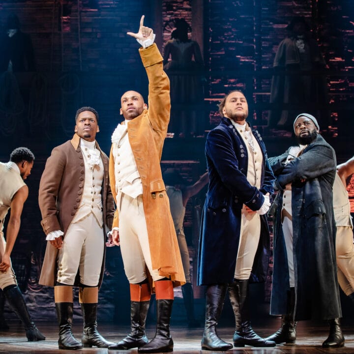Four men in period costumes stand at centre stage, one raising a hand, with a brick set and other actors in the background during a theatrical performance of Hamilton.