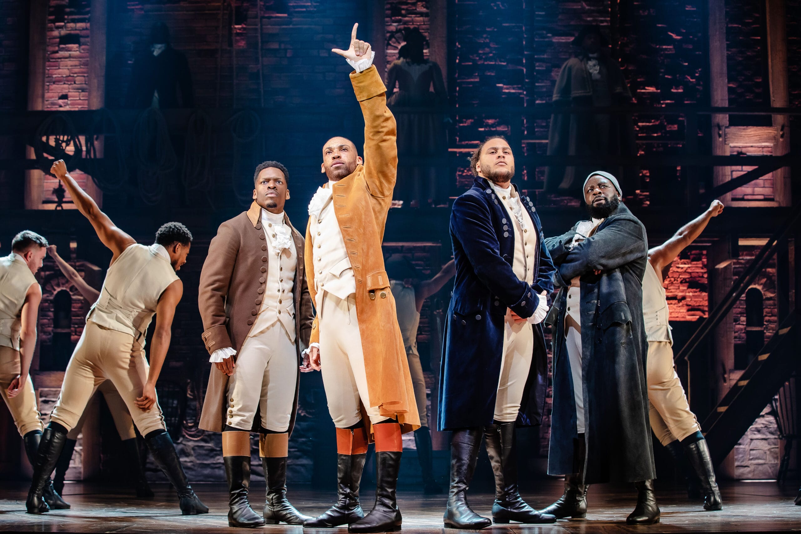 Four men in period costumes stand at centre stage, one raising a hand, with a brick set and other actors in the background during a theatrical performance of Hamilton.