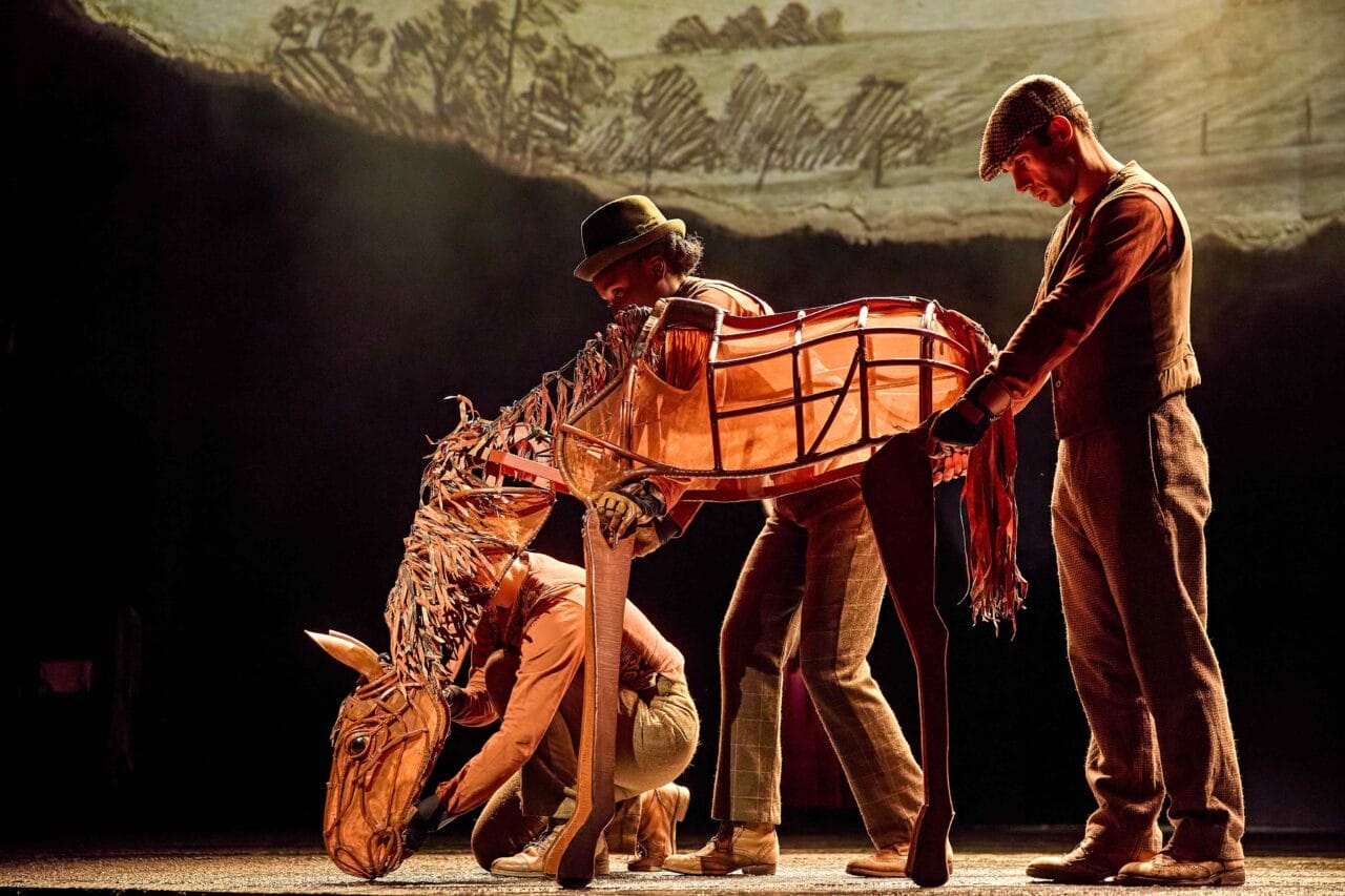 Three performers operate a large horse puppet onstage during a theatrical production, with a countryside backdrop in the background.
