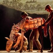 Three performers operate a large horse puppet onstage during a theatrical production, with a countryside backdrop in the background.