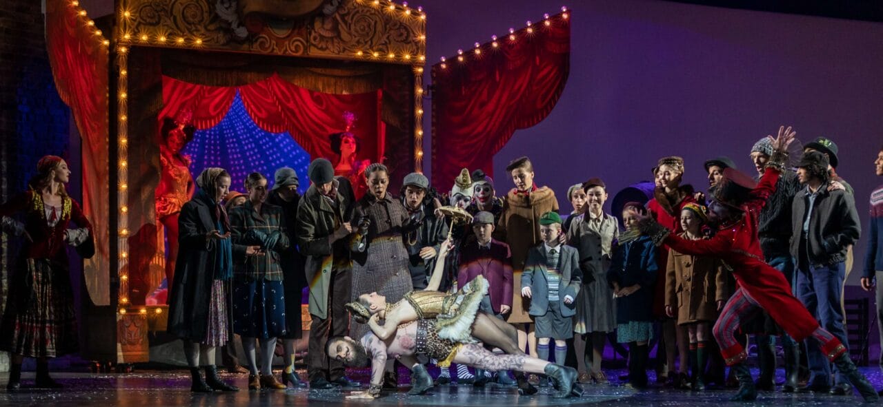 A theatrical stage scene features a large group of actors in period costumes, with two performers dramatically posed at center stage under bright lights.