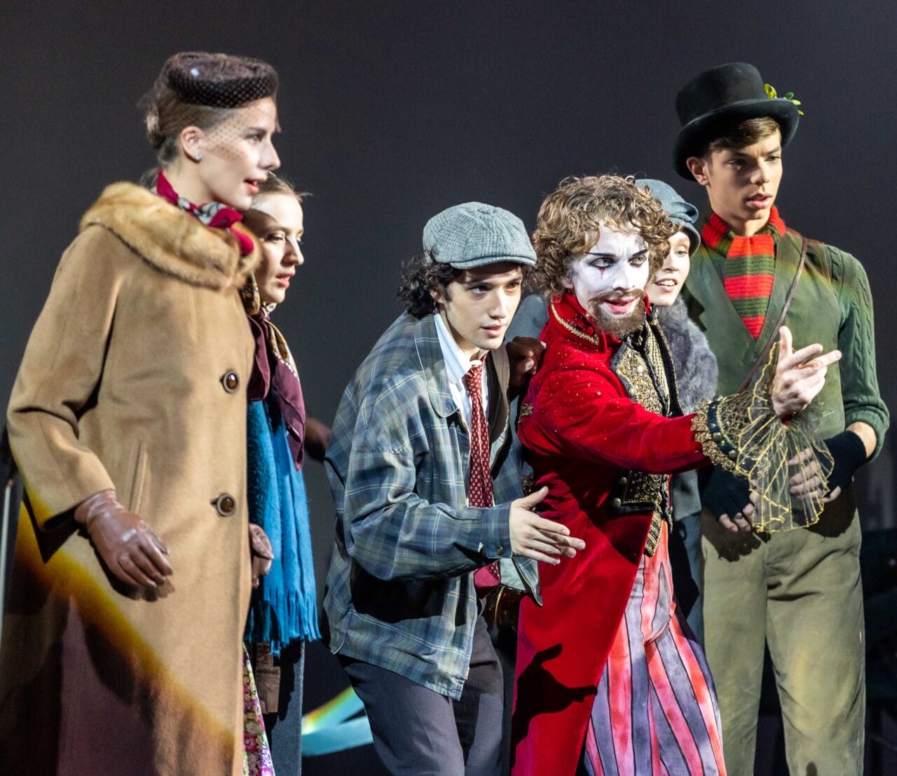 A group of six young people in theatrical costumes stand together on stage, with one person in white face paint and a red coat gesturing dramatically.