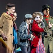 A group of six young people in theatrical costumes stand together on stage, with one person in white face paint and a red coat gesturing dramatically.