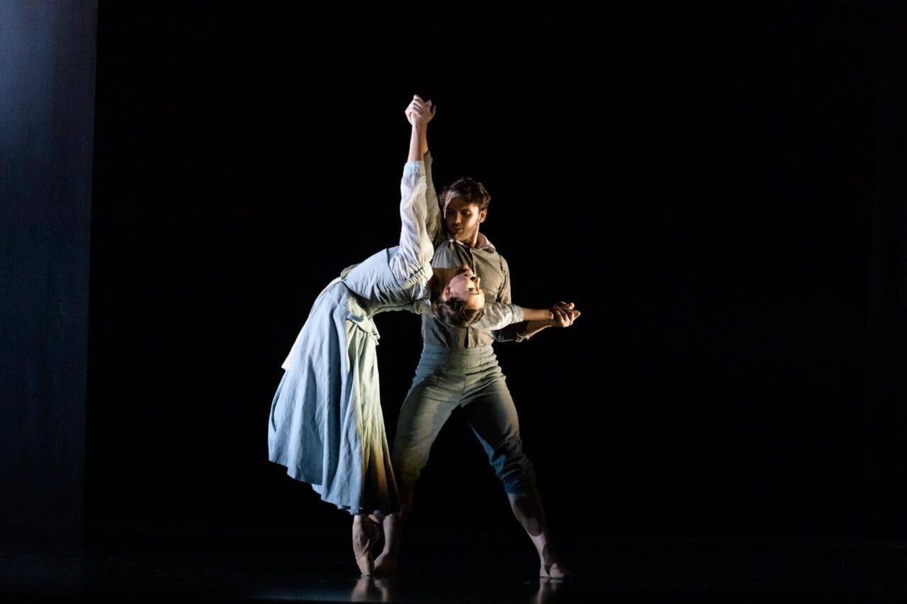 Two dancers perform a duet on stage; one leans back gracefully while holding the other's hand, both wearing light-colored costumes against a dark background.