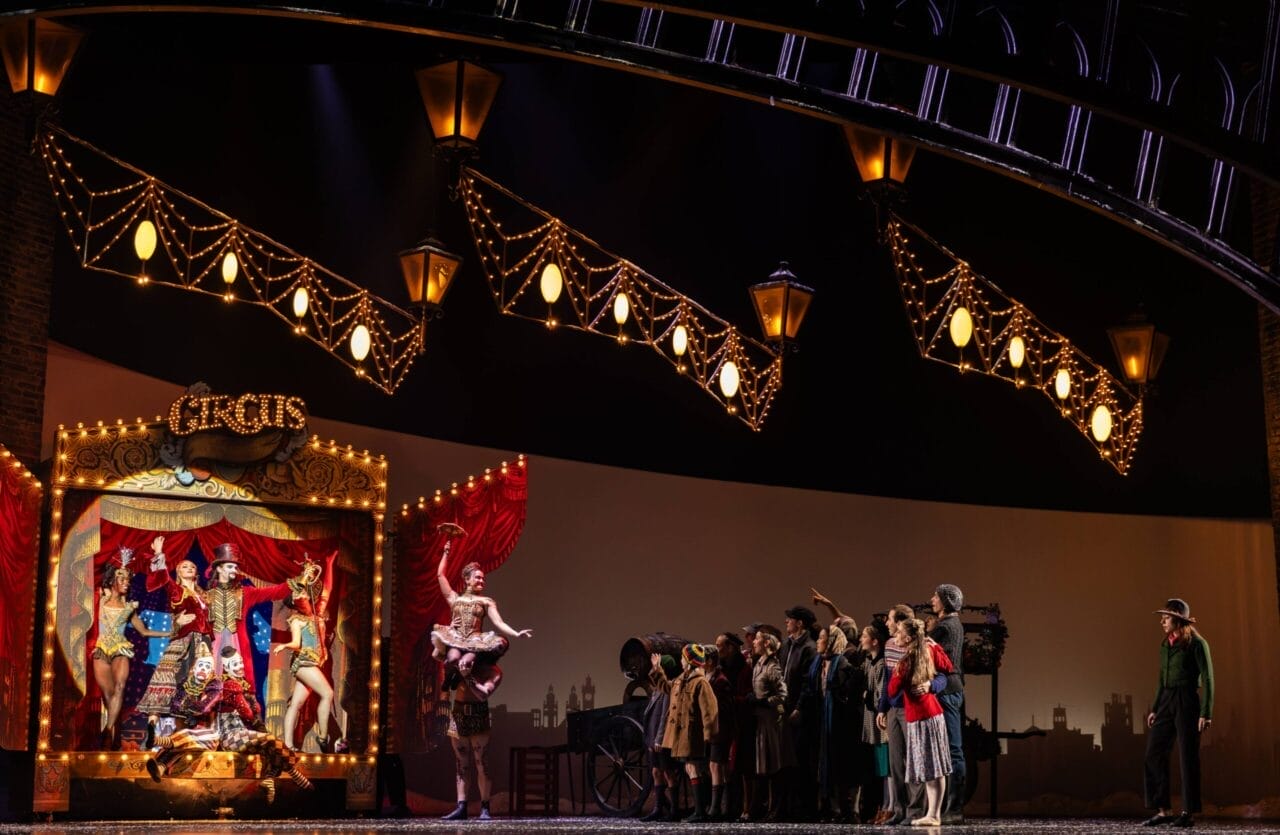 A stage scene features circus performers in colorful costumes posing on a lit circus booth, while the Snow Queen stands gracefully nearby, watched by a group of people under illuminated hanging lights.