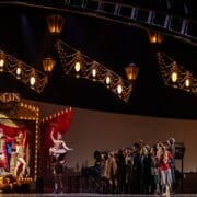 A stage scene features circus performers in colorful costumes posing on a lit circus booth, while the Snow Queen stands gracefully nearby, watched by a group of people under illuminated hanging lights.
