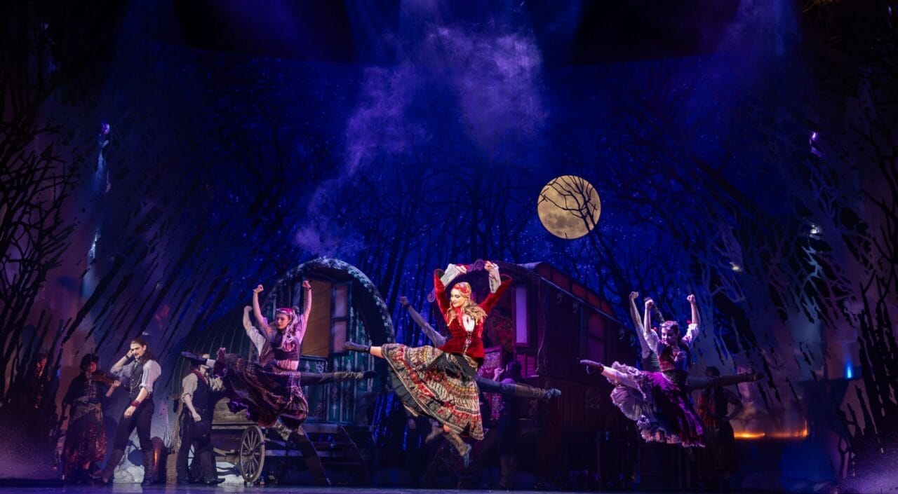 Performers in colorful costumes dance energetically on stage with a rural backdrop, trees, and a full moon, under dramatic lighting in this vibrant adaptation of Hans Christian Andersen’s beloved fairy tale, Snow Queen.