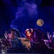 Performers in colorful costumes dance energetically on stage with a rural backdrop, trees, and a full moon, under dramatic lighting in this vibrant adaptation of Hans Christian Andersen’s beloved fairy tale, Snow Queen.