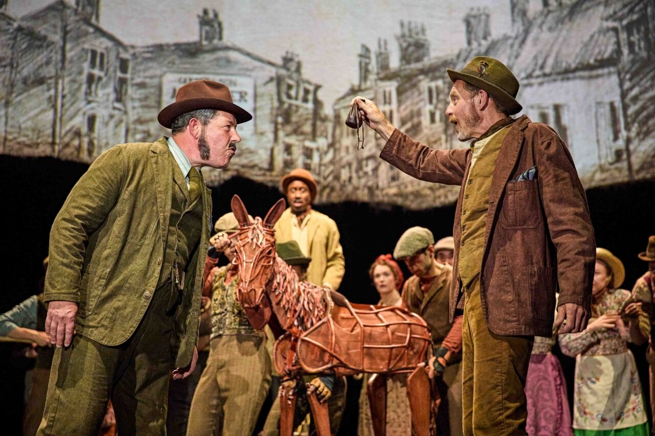 Two men in period costumes argue on stage, with one holding up an object; a group of actors and a horse puppet stand behind them, with a painted town backdrop.