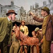 Two men in period costumes argue on stage, with one holding up an object; a group of actors and a horse puppet stand behind them, with a painted town backdrop.