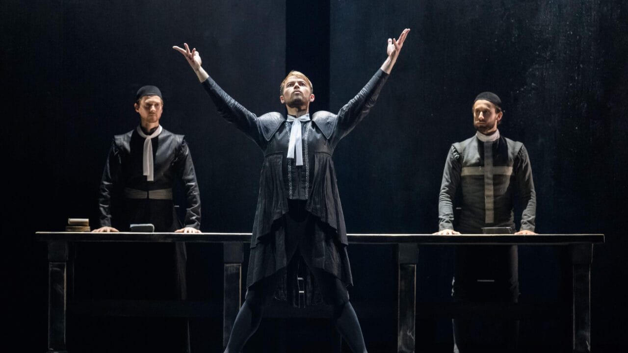 A theatrical performance shows three actors in dark costumes on stage. The central actor stands with raised arms, while the other two stand behind a table.