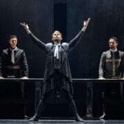 A theatrical performance shows three actors in dark costumes on stage. The central actor stands with raised arms, while the other two stand behind a table.
