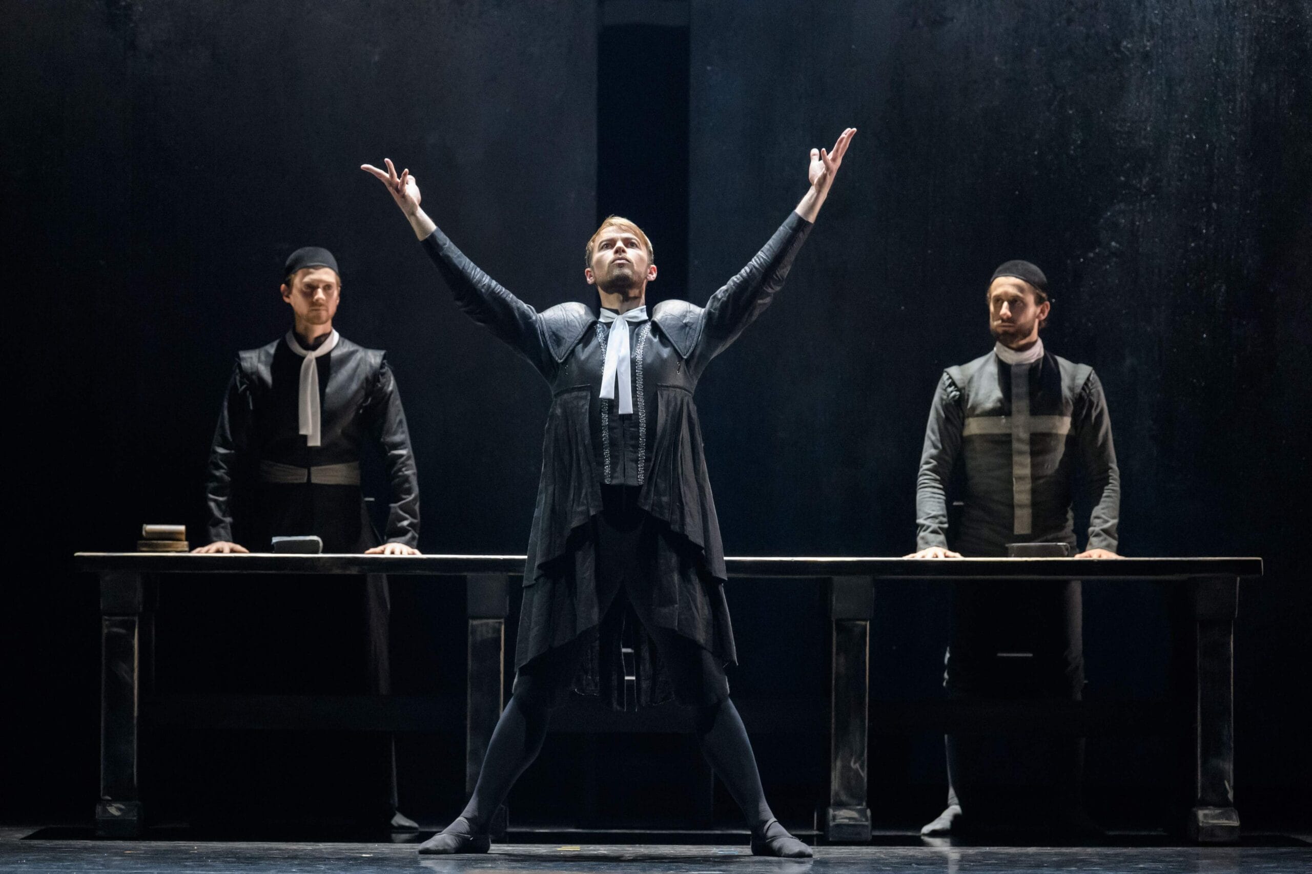 A theatrical performance shows three actors in dark costumes on stage. The central actor stands with raised arms, while the other two stand behind a table.
