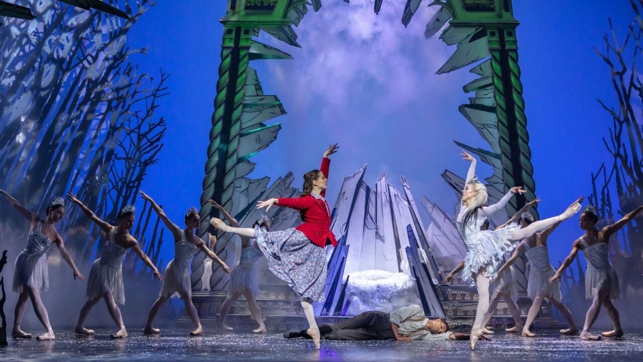 Ballet dancers perform on stage in front of a dramatic icy backdrop; one dancer wears red, others wear white, with one person lying on the ground.