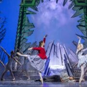 Ballet dancers perform on stage in front of a dramatic icy backdrop; one dancer wears red, others wear white, with one person lying on the ground.