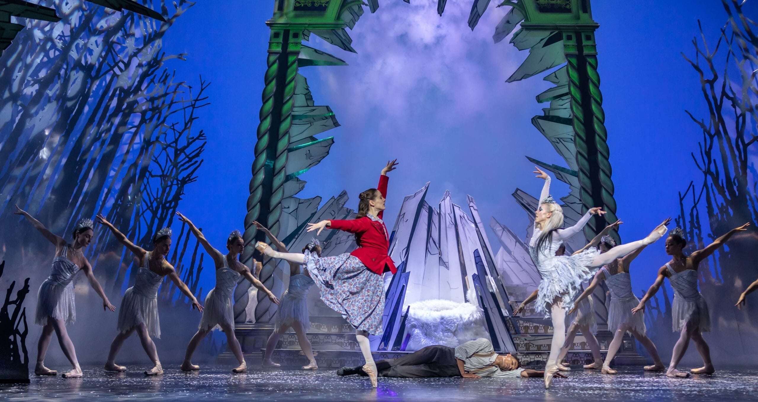 Ballet dancers perform on stage in front of a dramatic icy backdrop; one dancer wears red, others wear white, with one person lying on the ground.