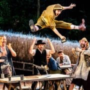 A group of men in period costumes watch as one man leaps high above a table with arms outstretched, mid-performance in an outdoor theatrical play.
