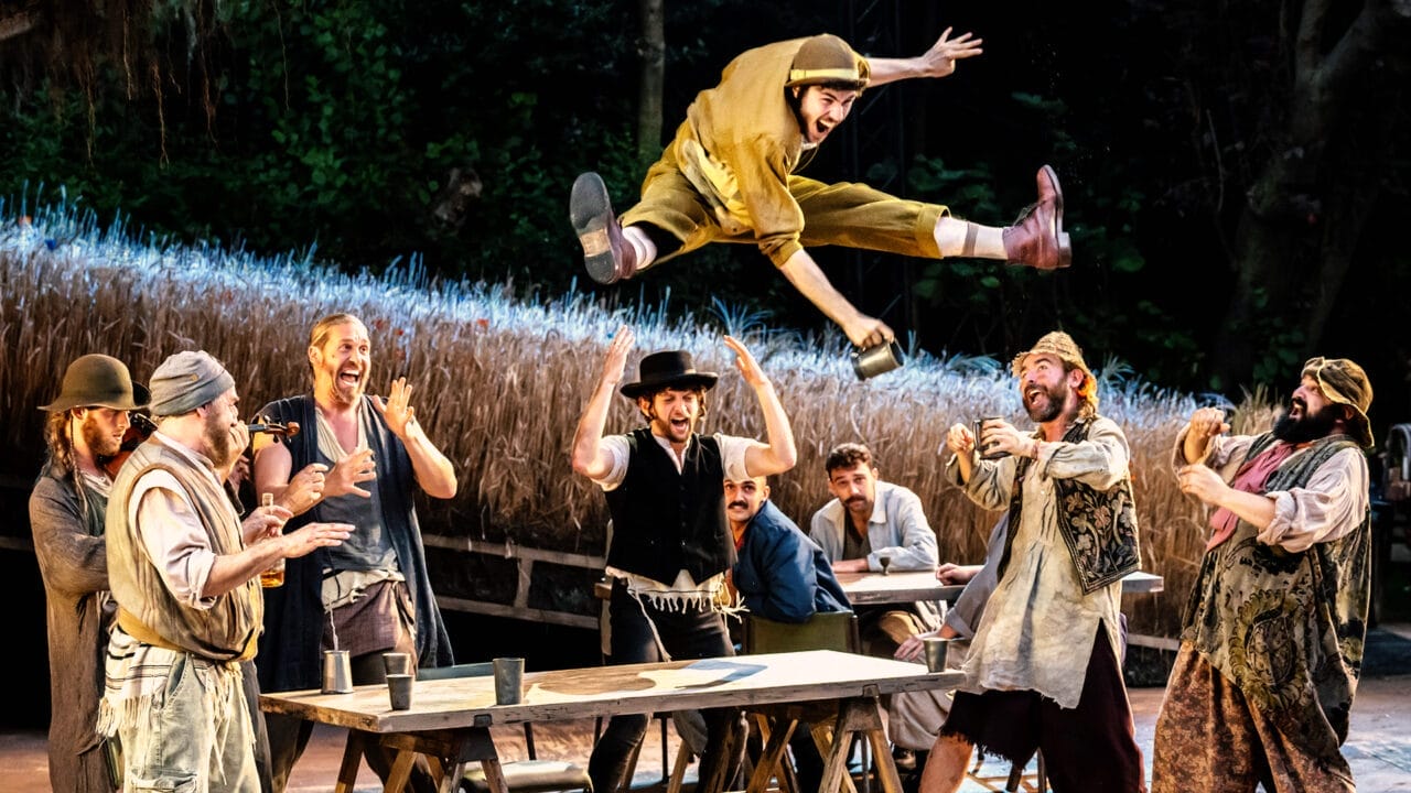 A group of performers in rustic costumes gathers around a table outdoors as one man leaps energetically mid-air above them, holding a metal cup, reminiscent of a lively scene from Fiddler on the Roof, while others watch and cheer.