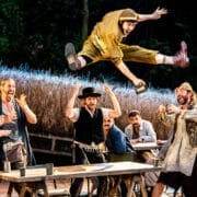 A group of performers in rustic costumes gathers around a table outdoors as one man leaps energetically mid-air above them, holding a metal cup, reminiscent of a lively scene from Fiddler on the Roof, while others watch and cheer.