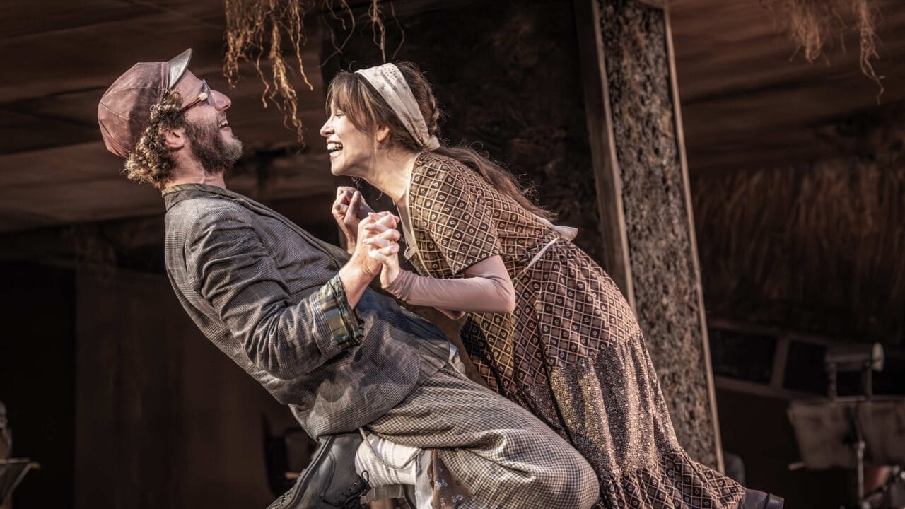 Two actors in period costumes hold hands and smile at each other while kneeling on a wooden table during a stage performance.