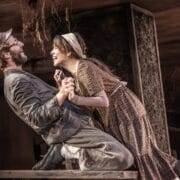 Two actors in period costumes hold hands and smile at each other while kneeling on a wooden table during a stage performance.