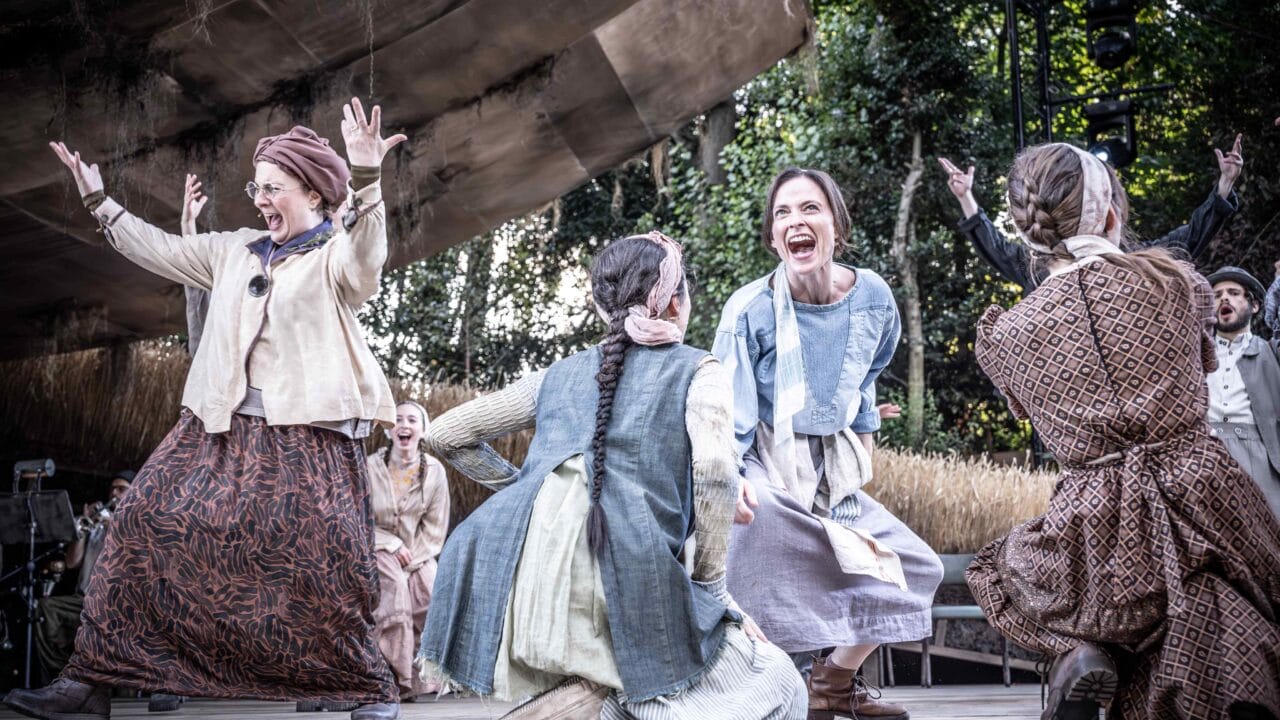 A group of women in period costumes celebrate energetically on an outdoor stage, surrounded by trees and rustic scenery.
