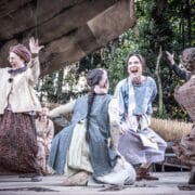 A group of women in period costumes celebrate energetically on an outdoor stage, surrounded by trees and rustic scenery.
