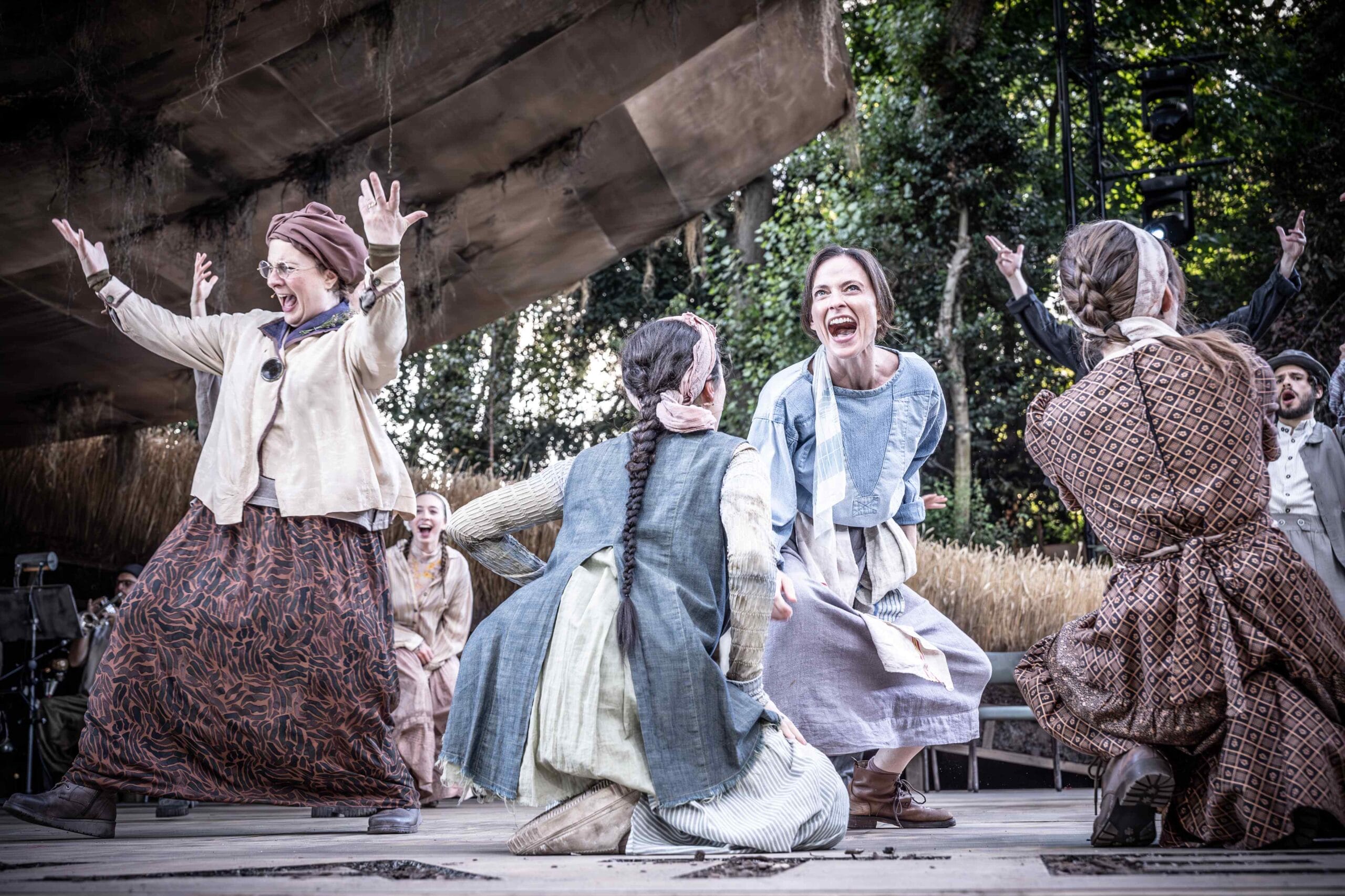 A group of women in period costumes celebrate energetically on an outdoor stage, surrounded by trees and rustic scenery.