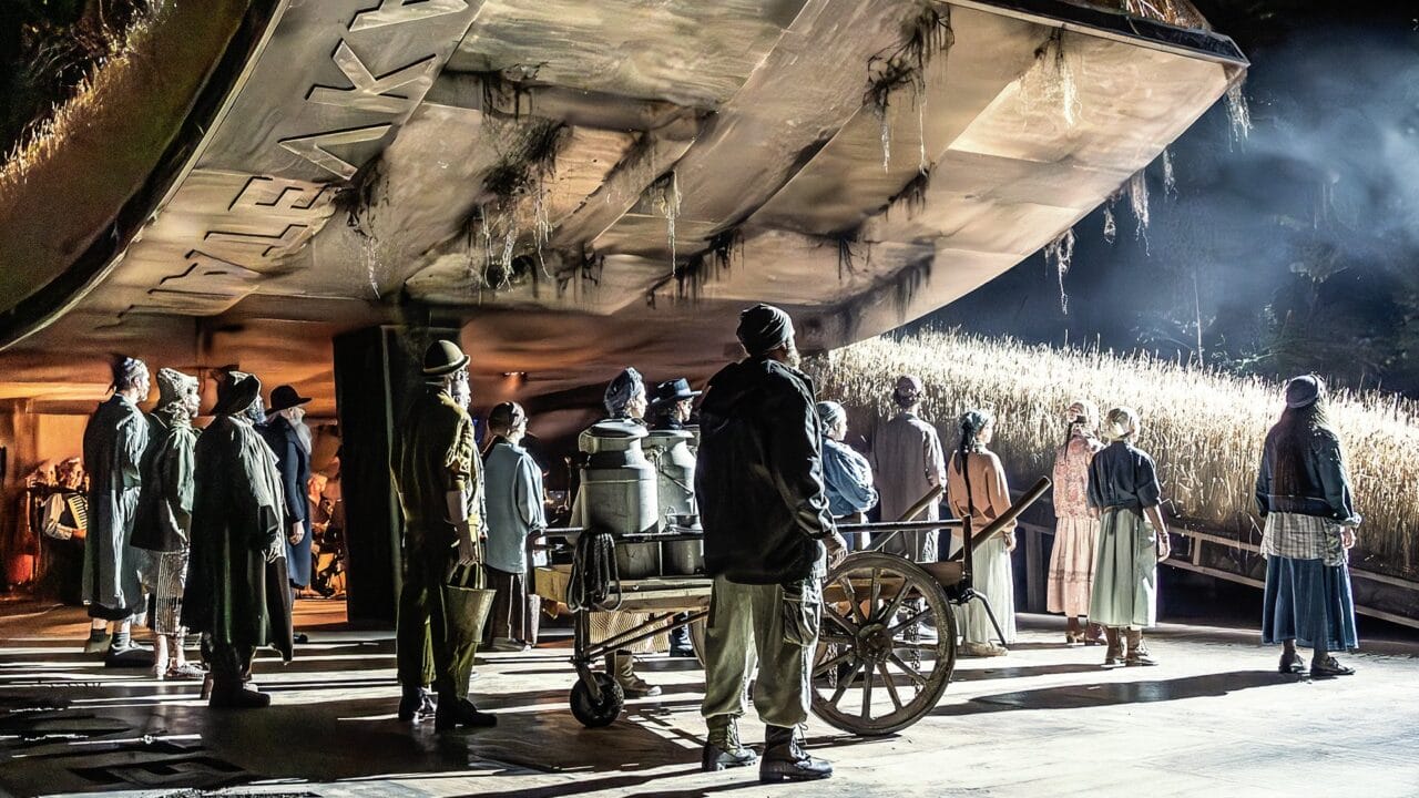 A group of people in period clothing stand under a large, weathered building beside a field of tall plants, with one person pushing a cart in the foreground.