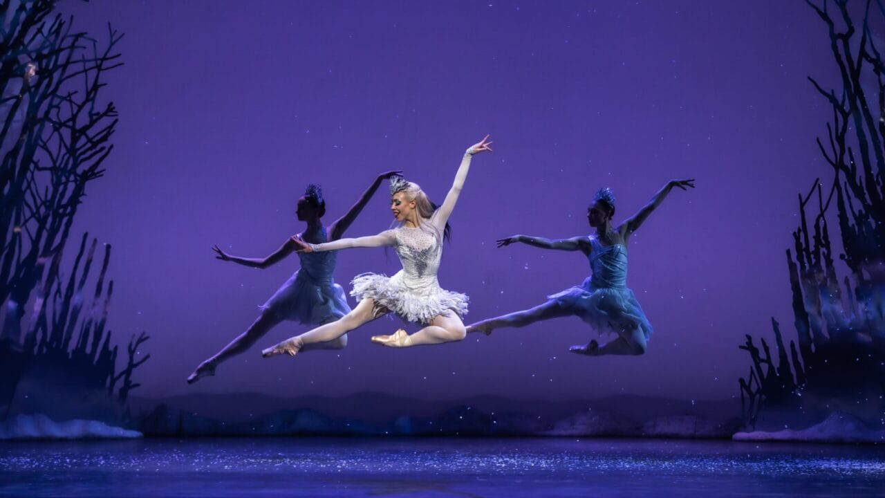 A ballerina in a white tutu performs a mid-air leap, flanked by two other dancers in blue costumes, against a purple background with silhouette trees.