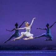 A ballerina in a white tutu performs a mid-air leap, flanked by two other dancers in blue costumes, against a purple background with silhouette trees.