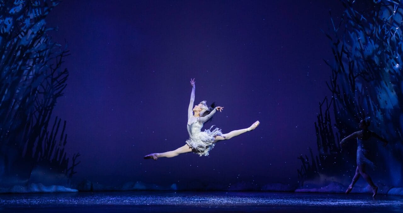 A ballet dancer in a white costume performs a mid-air split leap on stage, with a dark blue background and winter scenery.