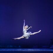 A ballet dancer in a white costume performs a mid-air split leap on stage, with a dark blue background and winter scenery.