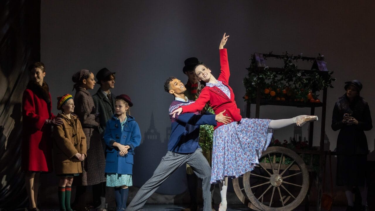 A group of people watch as a male and female ballet dancer perform in front of a flower cart on stage, with the woman posed en pointe and the man supporting her.