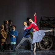 A group of people watch as a male and female ballet dancer perform in front of a flower cart on stage, with the woman posed en pointe and the man supporting her.