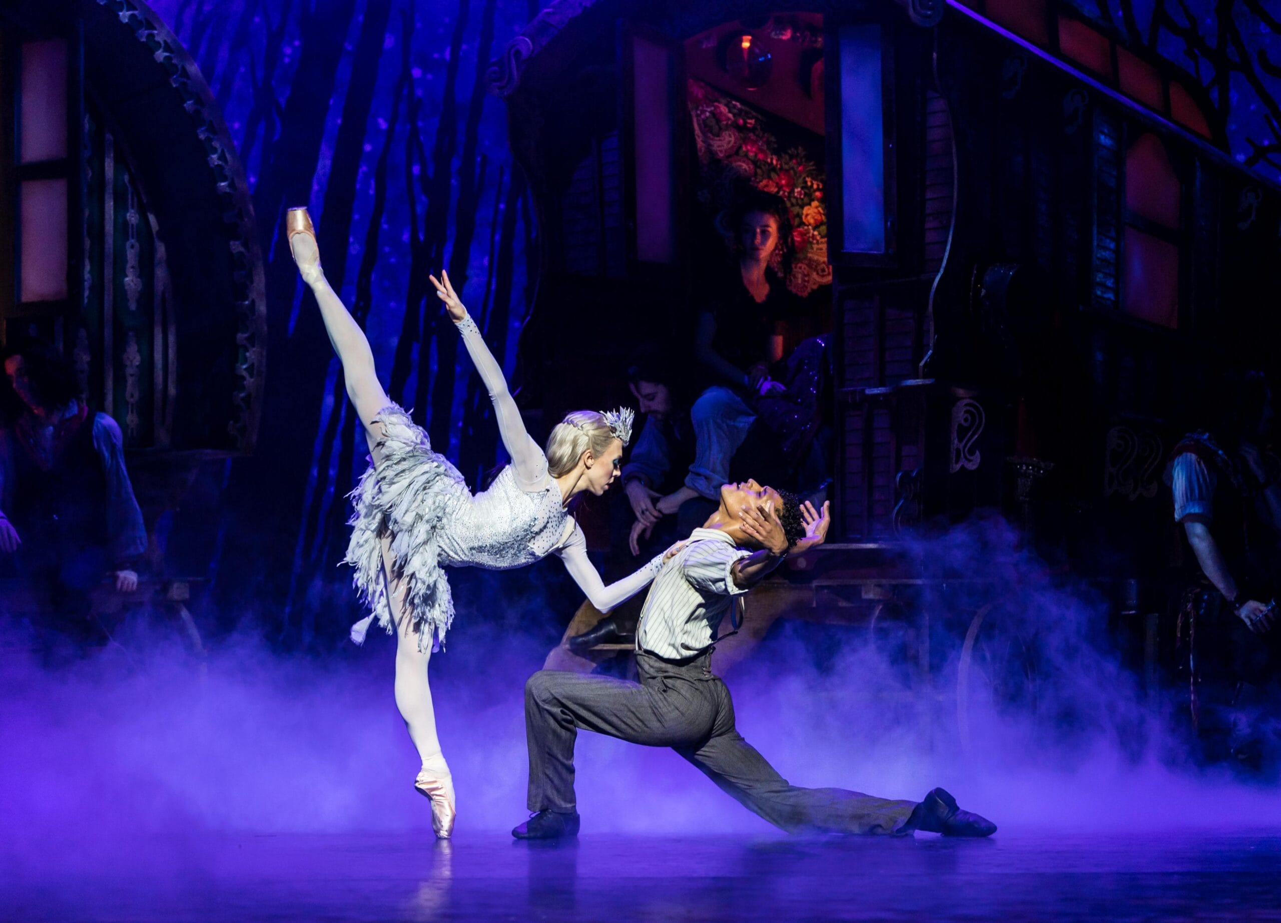 A ballet dancer in a white costume performs an arabesque, leaning toward a kneeling male dancer. They are on a theatrical stage with a misty forest backdrop.