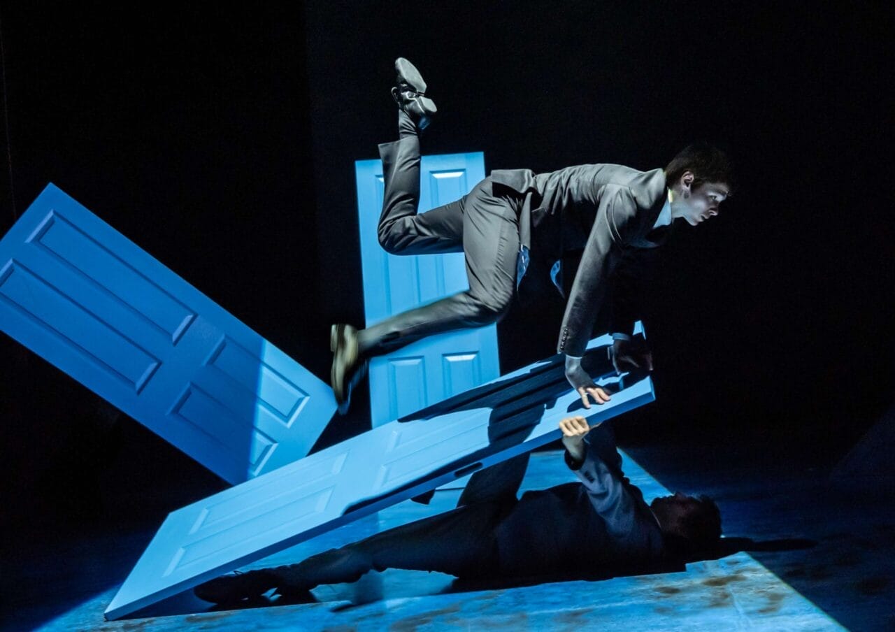 Two people in suits perform acrobatic moves with three blue doors on a dark stage, evoking Mod Ballet style—one jumping over a door while the other lies on the ground holding a door up, reminiscent of Quadrophenia’s bold energy.